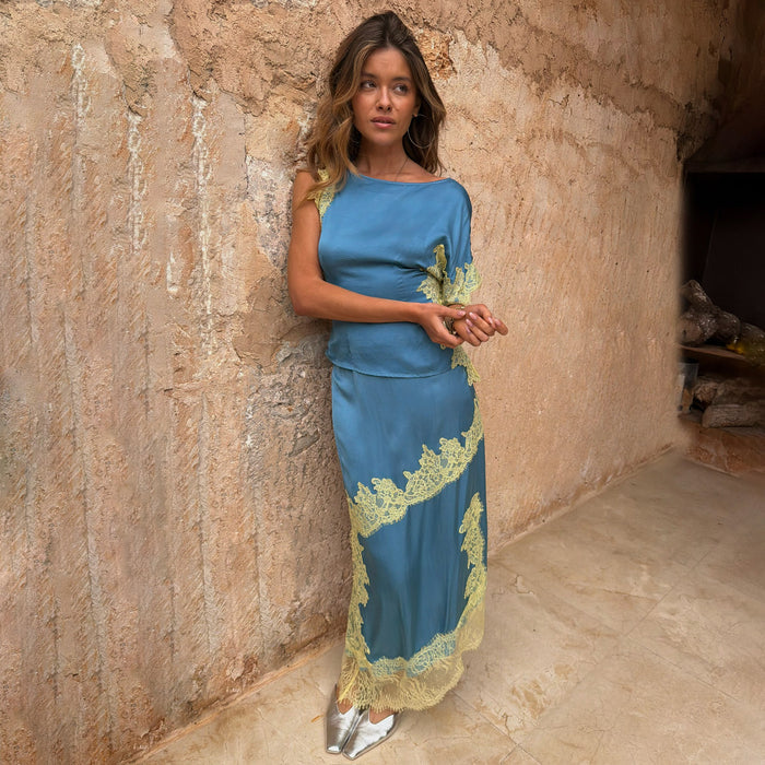 Blue and Lemon Millie Skirt and one-shoulder top co-ord with lace trim, leaning against a stone wall.
