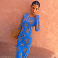 Blue Eye Mesh Dress with eye and heart motifs, a long-sleeved fitted maxi silhouette and tan clutch for vacation.