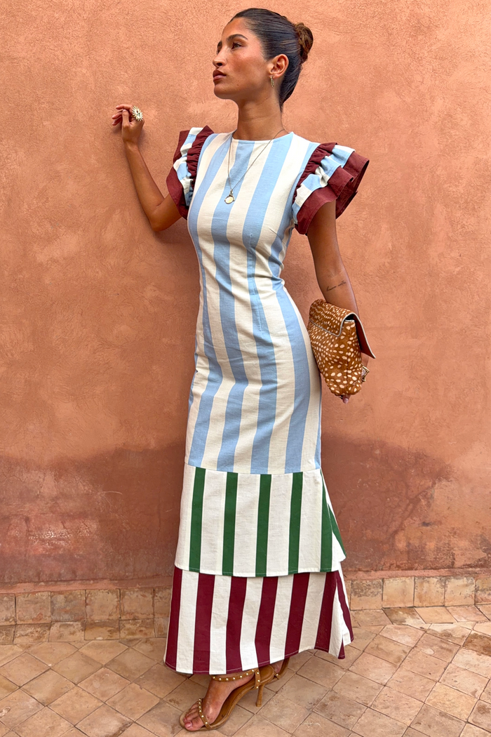 Blue Stripe Alena Midi Dress in cotton-linen with burgundy ruffle sleeves and a tiered hem against a terracotta wall.