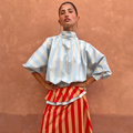 Blue Stripe Batwing Blouse with a high neck and shirred cuffs, worn by a model against a terracotta wall.