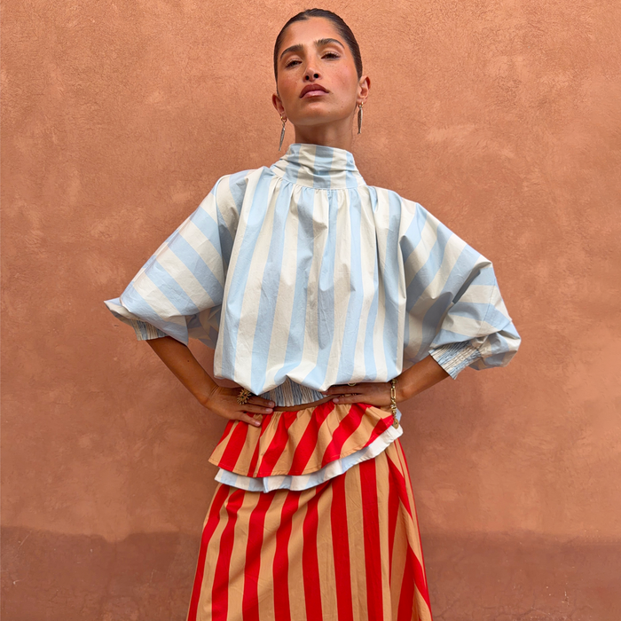 Blue Stripe Batwing Blouse with a high neck and shirred cuffs, worn by a model against a terracotta wall.