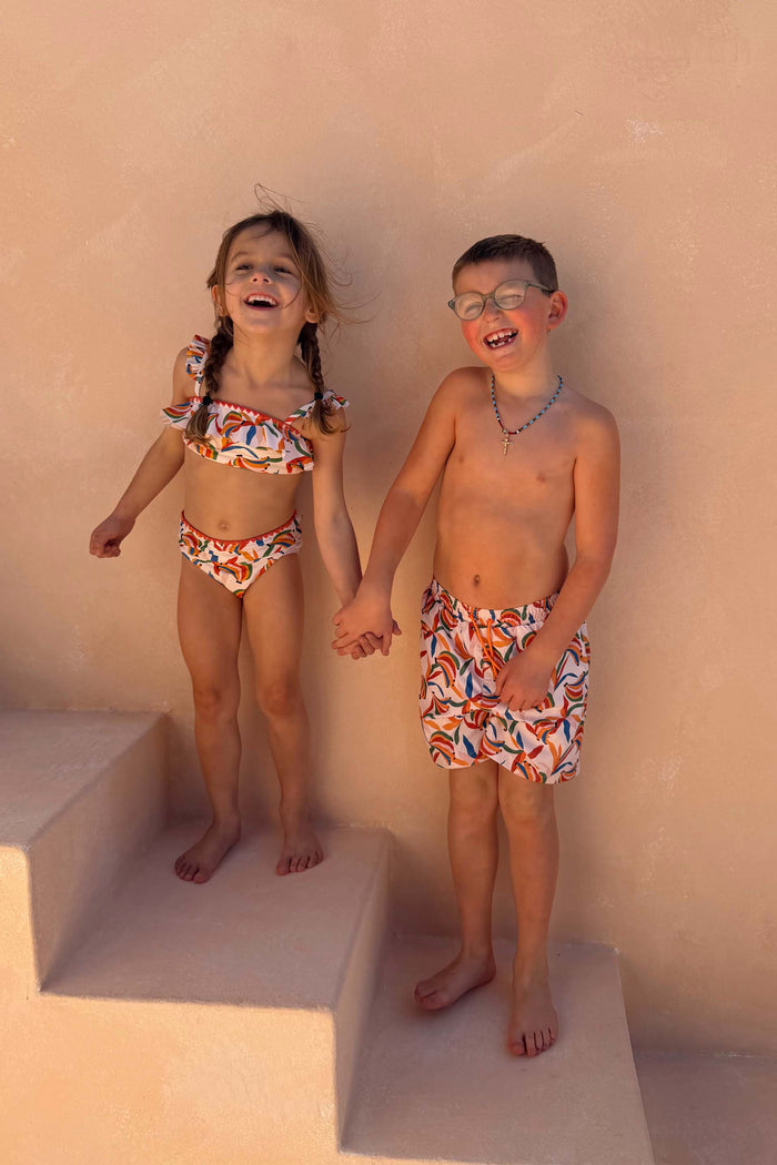 Boy's Banana Print Swim Shorts in cream with a multi-coloured print, worn by a child laughing on stairs.