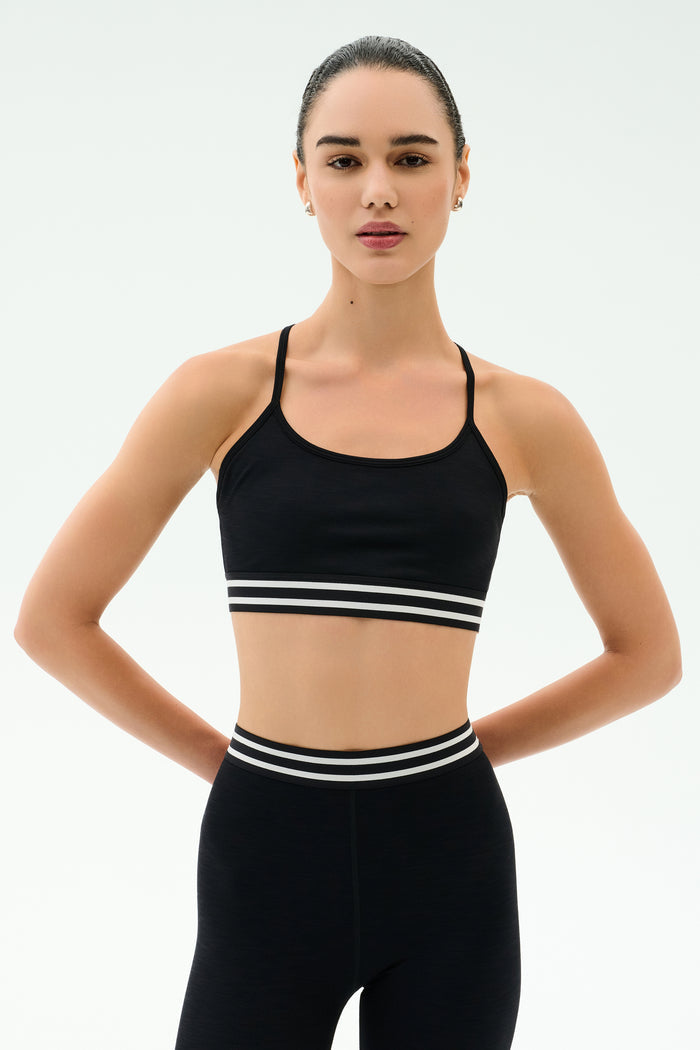 A woman with dark hair tied back wears the SPLITS59 Cali Active Rib Bra in black and matching high-waisted leggings, standing against a plain white background with her hands behind her back.