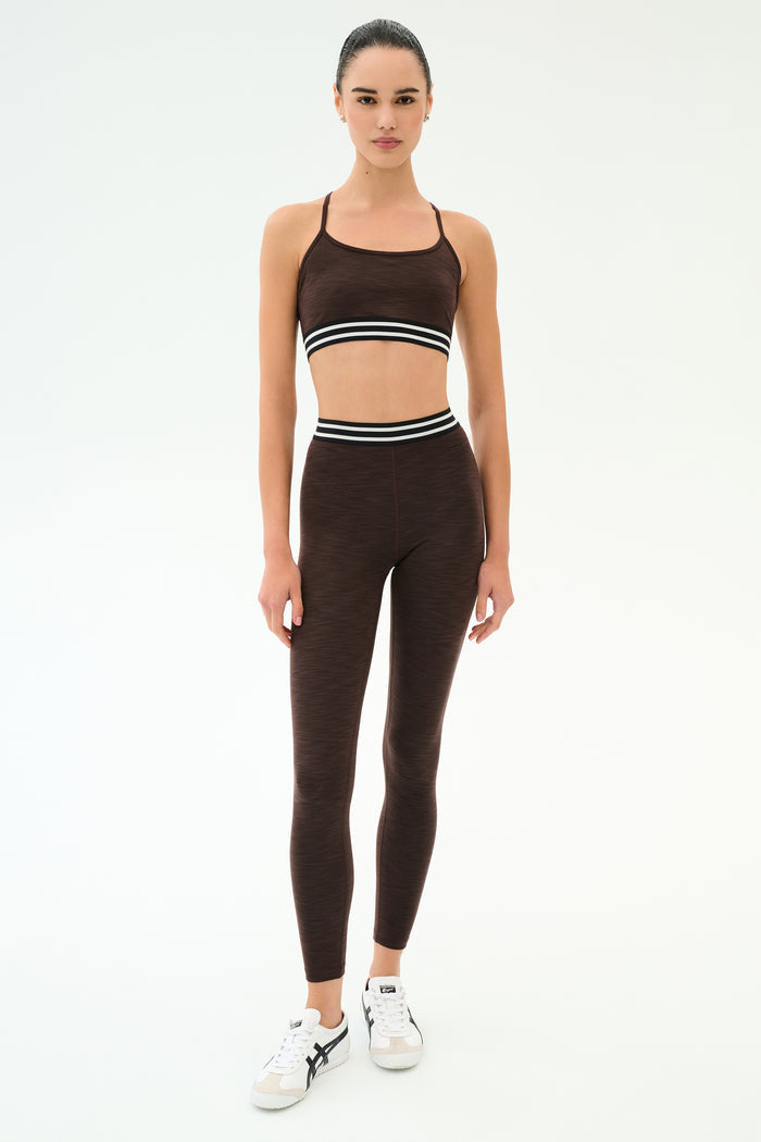 A woman wears the SPLITS59 Cali Active Rib Bra in Heather Dark Chocolate with matching leggings, featuring white and black striped waistbands, standing against a plain white background.