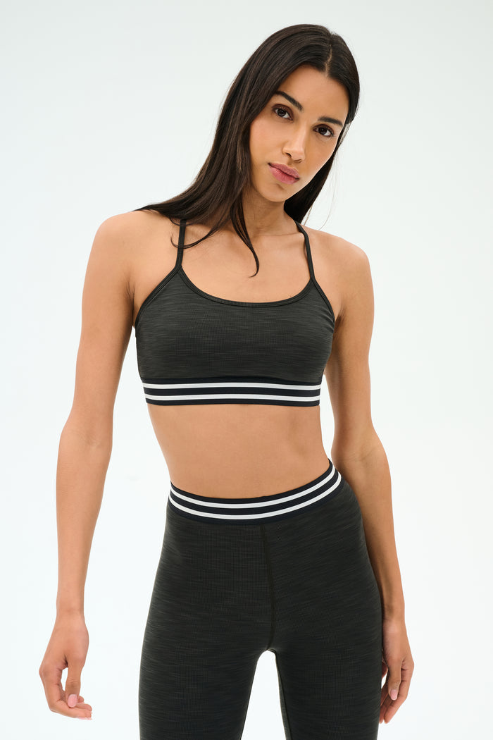 A person models the SPLITS59 Cali Active Rib Bra in Heather Graphite, featuring a supportive dark design, against a plain light background.