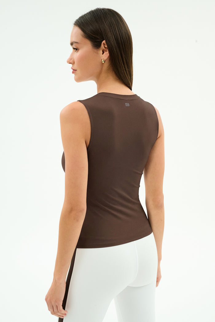 A woman with straight dark hair wears the SPLITS59 Carter Airweight Lite Tank in Dark Chocolate and white pants, facing away in a minimal studio. The tank’s buttery soft fabric adds comfort to her simple look.
