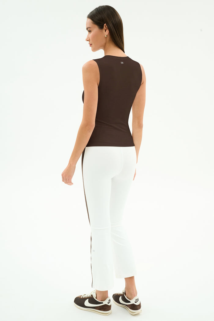A woman faces away on a plain white background, wearing the SPLITS59 Carter Airweight Lite Tank in Dark Chocolate, paired with white pants featuring a brown stripe and brown Nike sneakers.