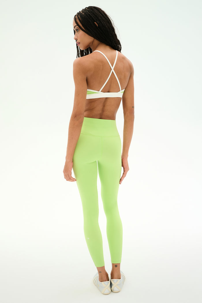 A person is standing in a studio facing away from the camera, wearing the SPLITS59 Clare High Waist Rigor 7/8 set in Limeade/White—a light green sports bra with white crisscrossed straps and matching leggings crafted from soft nylon spandex fabric—paired with slip-on shoes.