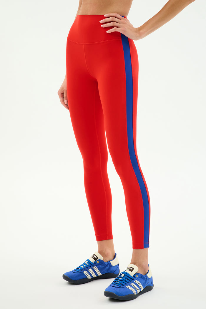Wearing SPLITS59’s Clare High Waist Rigor 7/8 leggings in Lotus Red/Cobalt, this person pairs performance fabric activewear with blue athletic shoes, standing against a white background—ready to work out.