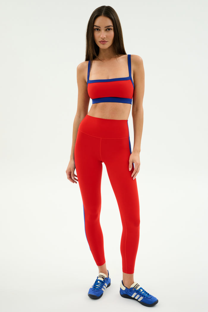 A woman wears the SPLITS59 Clare High Waist Rigor 7/8 leggings in Lotus Red/Cobalt, featuring performance fabric for workout comfort, paired with blue athletic shoes, and stands against a plain white background.