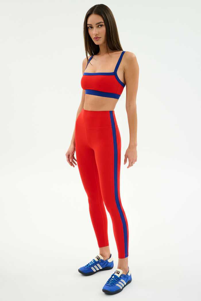 A woman wears the SPLITS59 Clare High Waist Rigor 7/8 leggings in Lotus Red/Cobalt with a matching crop top and blue sneakers, standing against a white background—ideal athletic style for your next workout.