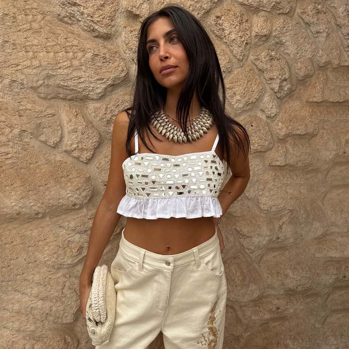 Cream Mirror Gabbi Top with mirror embellishments and a ruffle hem, styled with trousers against a stone wall.