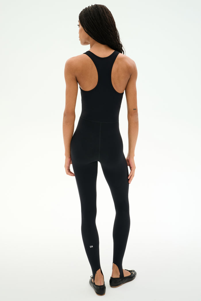 A woman faces away in the SPLITS59 Darci Airweight Stirrup Jumpsuit - Black, a fitted, stretch black sleeveless jumpsuit paired with black ballet flats, set against a plain white background.