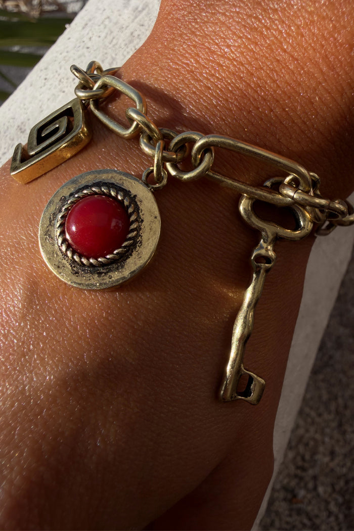 Brushed gold Deloris Charm Bracelet featuring a red stone pendant and key charm on a chunky chain, shown on a wrist.