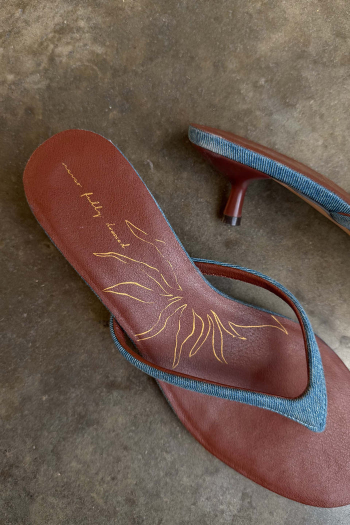 Denim Sandal Mule featuring slim denim straps, a brown cushioned sole with a gold sun motif and sculpted kitten heel.