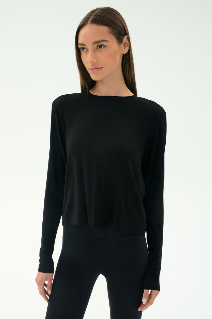A woman wears the SPLITS59 Djuna Long Sleeve in black, paired with black leggings and standing against a plain light background—ideal women's activewear for cool weather workouts.