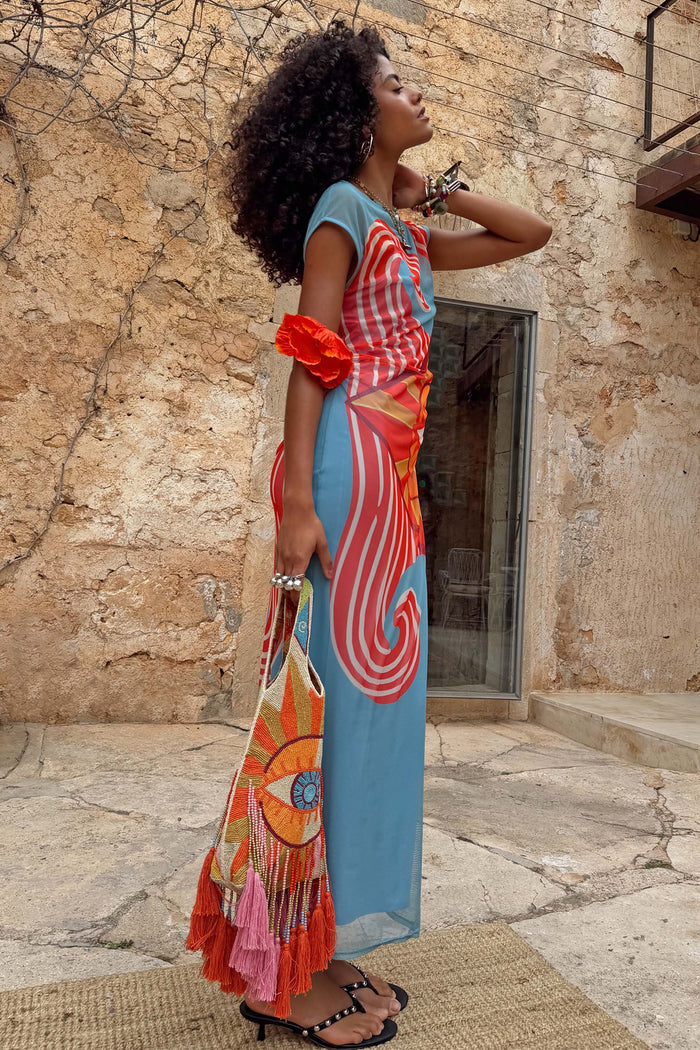 Blue Dolce Vita Mesh Dress with vibrant red swirl motifs, styled with a beaded eye bag against a stone wall.