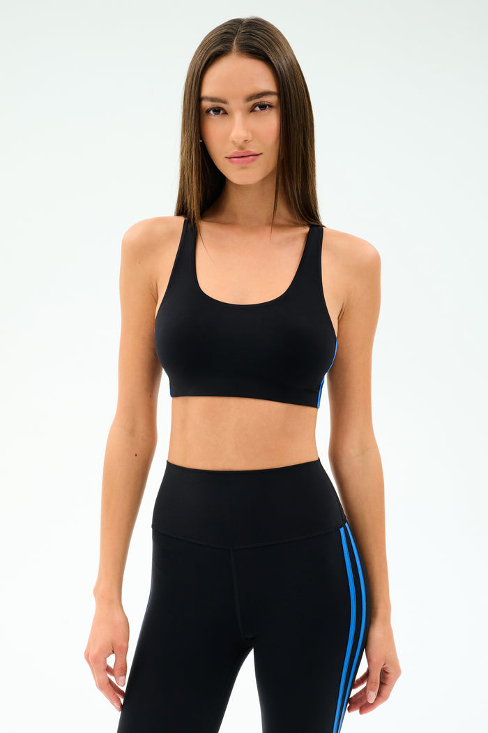 A woman stands against a plain background, wearing SPLITS59’s Ella Airweight Bra in Black/Classic Blue, made from soft nylon spandex for comfort and flexibility, paired with matching black leggings featuring blue side stripes.