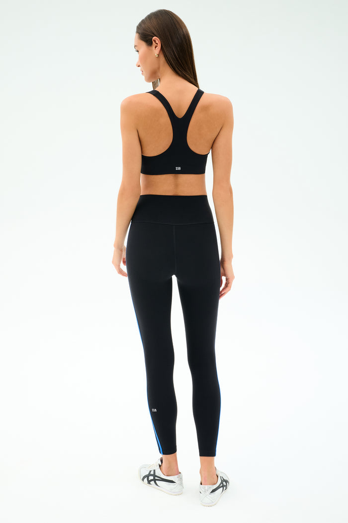 A woman faces away, wearing the SPLITS59 Ella Airweight Bra in Black/Classic Blue with high-waisted nylon spandex leggings and white sneakers, set against a plain white background.