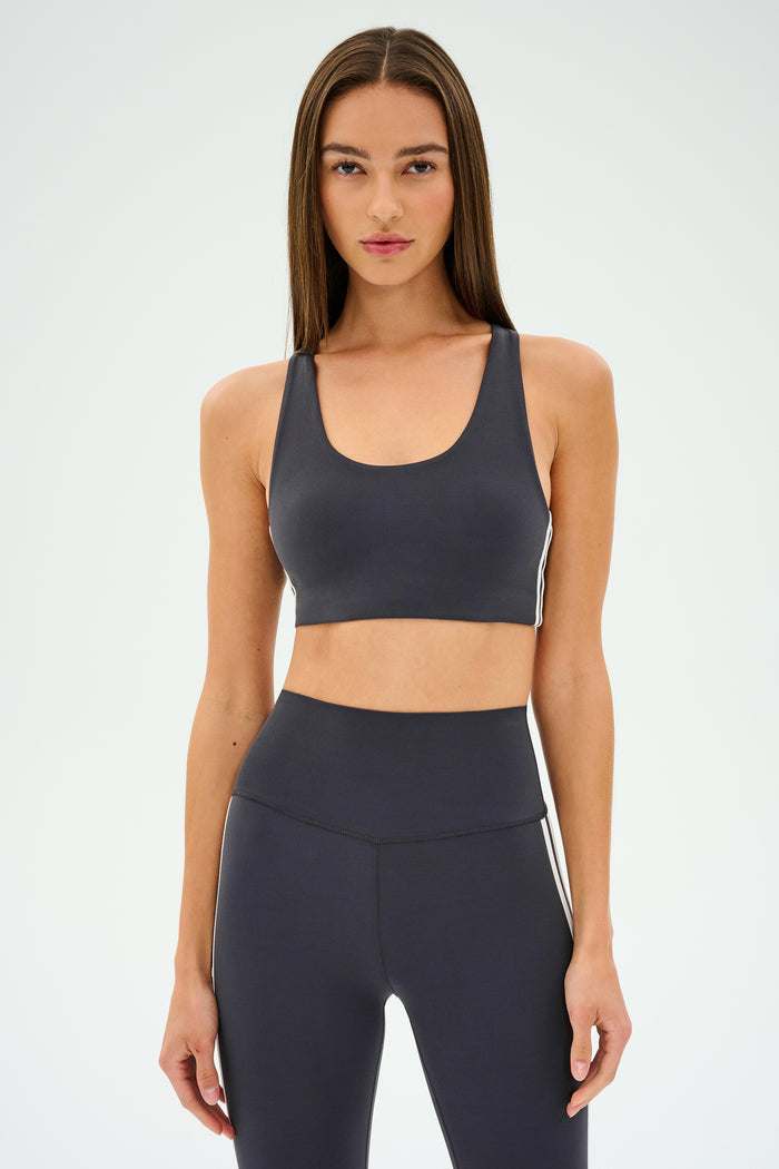 A person wearing an Ella Airweight Bra in Graphite/White from SPLITS59 stands against a plain background, ready for hot yoga.