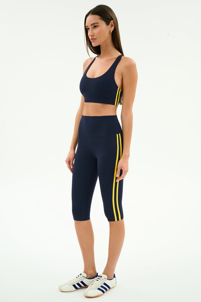 A woman wears the SPLITS59 Ella Airweight Bra in Indigo/Bright Yellow, featuring stretch nylon-spandex fabric and yellow side stripes, paired with white Adidas sneakers, posing against a plain white background.