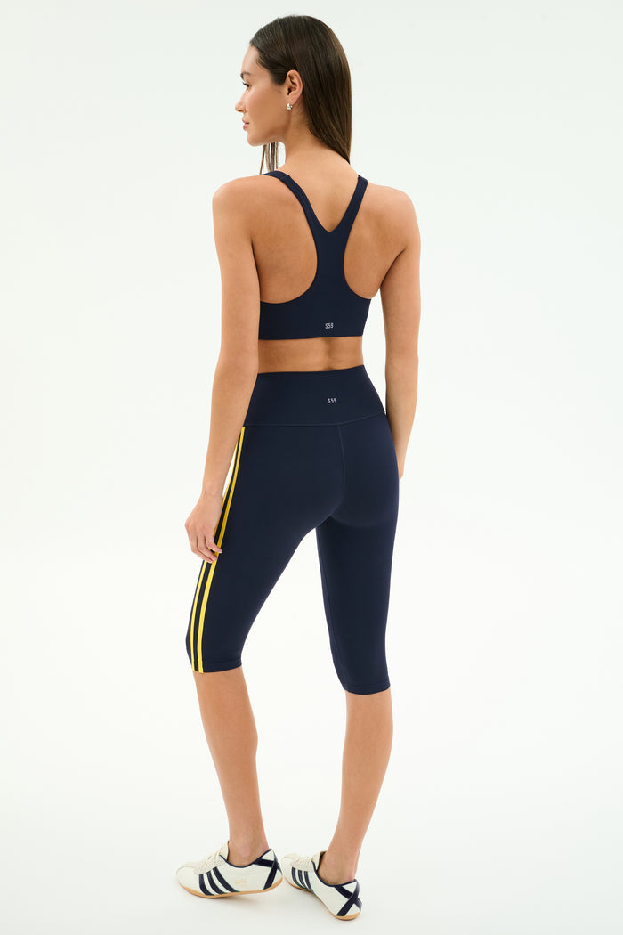 A woman stands facing away, wearing the SPLITS59 Ella Airweight Bra in Indigo/Bright Yellow and matching capri leggings, paired with white sneakers with black stripes, set against a plain background for an activewear look.