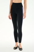 A person wears SPLITS59 Ella High Waist Airweight 7/8 leggings in Black/Purple Haze, featuring two side stripes, with black ballet flats, standing against a plain white background.