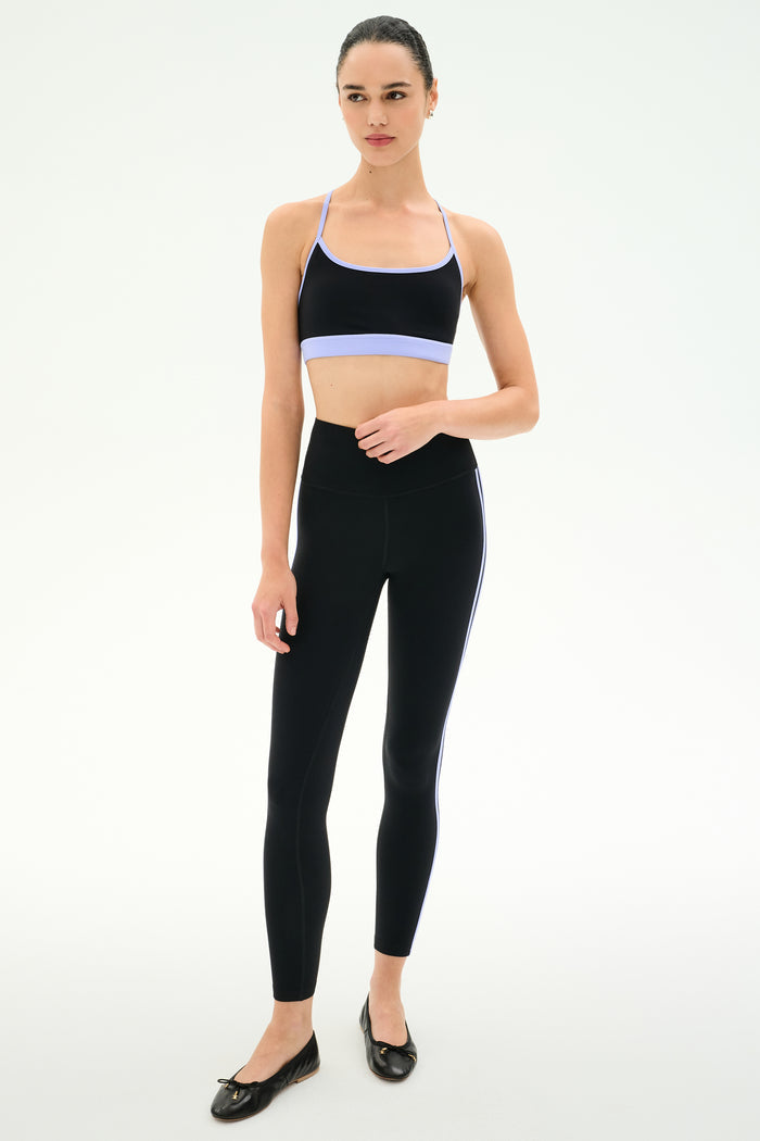 A woman stands against a plain background wearing SPLITS59's Ella High Waist Airweight 7/8 leggings in Black/Purple Haze, paired with a black sports bra and black ballet flats.