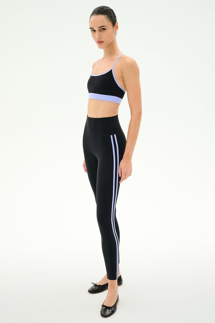 A woman models the SPLITS59 Ella High Waist Airweight 7/8 leggings in Black/Purple Haze with side stripes, paired with a black sports bra and black ballet flats, against a plain white background.