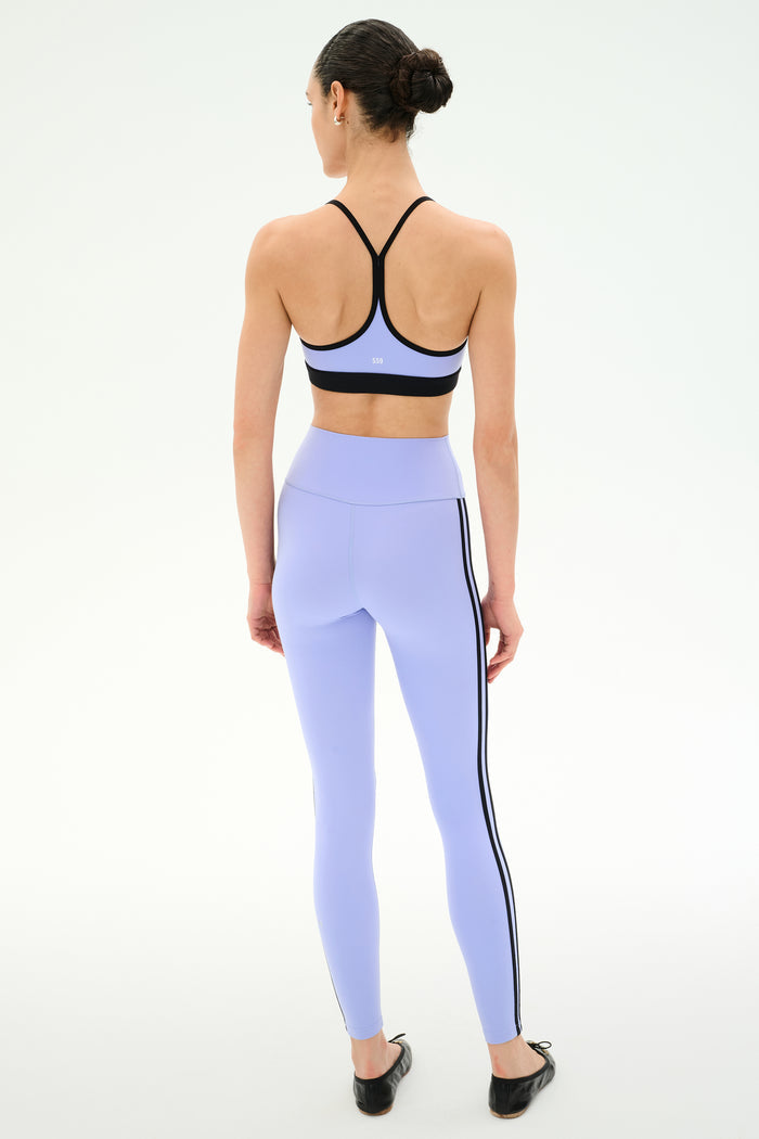 A woman stands with her back to the camera, wearing SPLITS59’s Ella High Waist Airweight 7/8 leggings in Purple Haze/Black, featuring black side stripes, set against a plain light background.