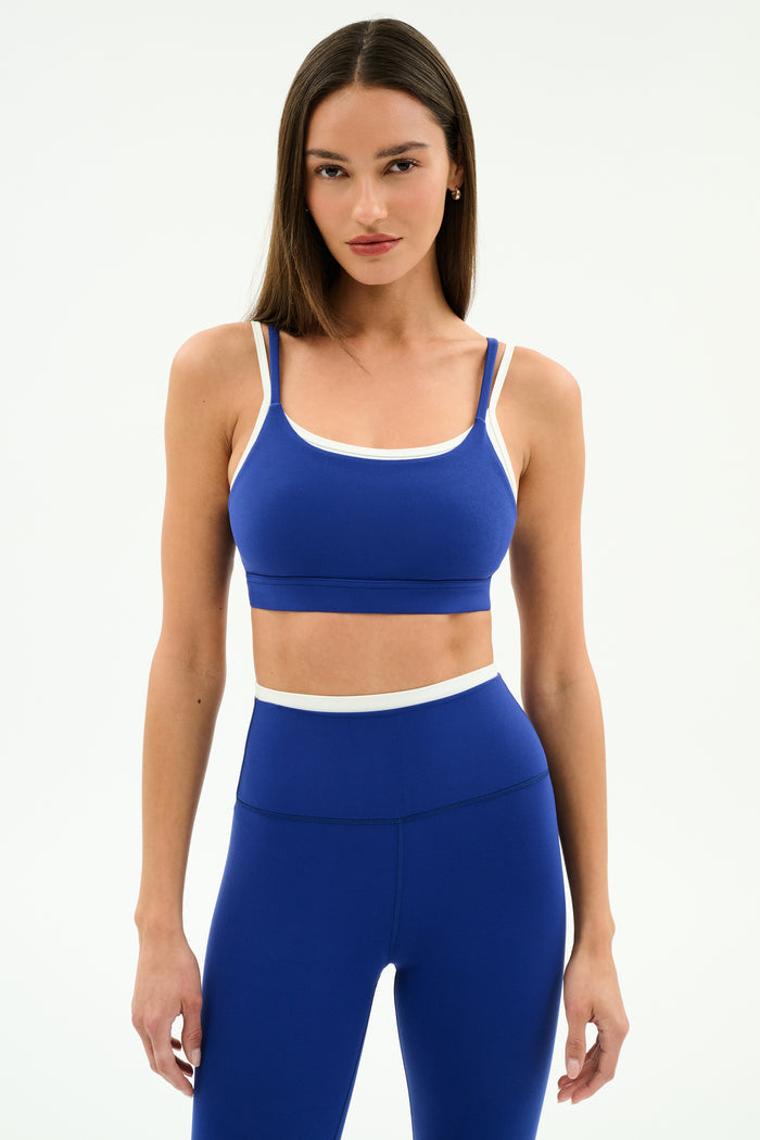 A woman poses against a plain background in the SPLITS59 Elliah Airweight Double Layer Bra in cobalt/white, styled with matching high-waisted leggings featuring white trim.