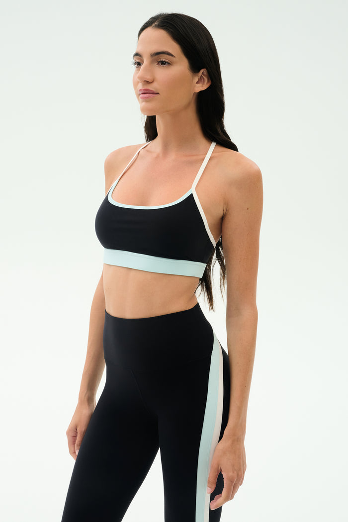 A woman stands in a studio wearing the SPLITS59 Emerson Airweight Bra in Black/Ice, featuring white and light blue trim, paired with high-waisted leggings—an ideal look for Pilates or hot yoga.