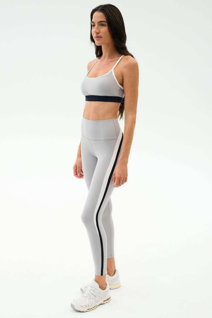 A woman wears the SPLITS59 Emerson Airweight Bra in Dove Grey/Indigo and matching high-waisted leggings, ideal for Pilates or hot yoga. She pairs them with white sneakers and stands against a plain white background.