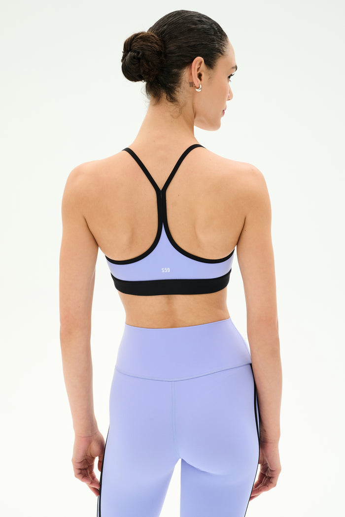 A person with dark hair in a bun is seen from behind wearing the SPLITS59 Emerson Airweight Bra in Purple Haze/Black and matching leggings.
