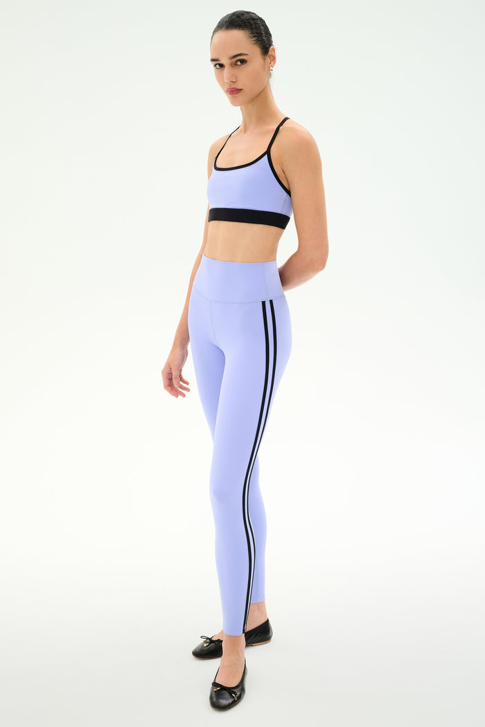 A person wears the SPLITS59 Emerson Airweight Bra in Purple Haze/Black, paired with matching high-waisted leggings featuring side stripes and black ballet flats.