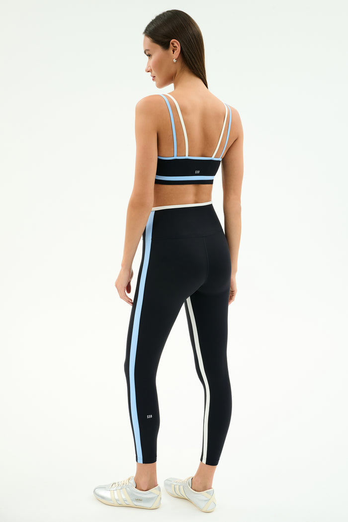 A woman stands in profile wearing the SPLITS59 Eva Rigor Double Strap Bra in Black/Skylight, paired with black leggings featuring blue and white stripes and white sneakers, set against a plain white background.