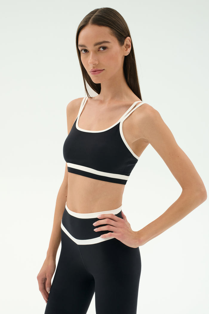 A woman wears the SPLITS59 Eva Rigor Double Strap Bra in Black/White, paired with matching high-waisted leggings, standing with one hand on her hip against a plain white background.