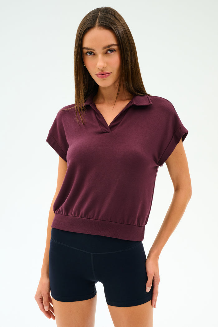 A woman faces forward, wearing the SPLITS59 Evie Fleece Sleeveless Polo in Dark Cherry and black shorts against a plain white background.