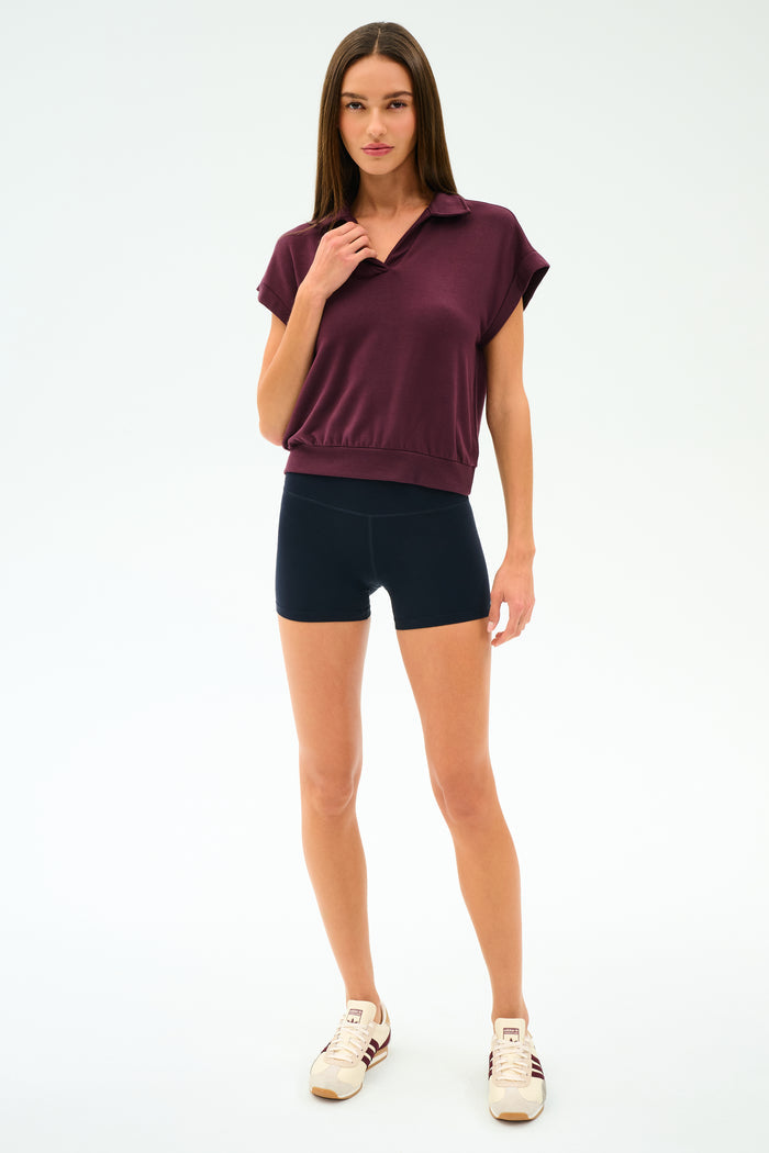 A woman with straight brown hair and a neutral expression stands against a white background wearing the SPLITS59 Evie Fleece Sleeveless Polo in Dark Cherry, paired with black shorts and white sneakers featuring maroon stripes.