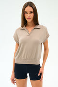 A woman poses against a plain background wearing the SPLITS59 Evie Fleece Sleeveless Polo in Latte and black fitted stretch shorts.