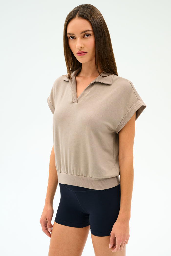 A woman with straight brown hair wears the SPLITS59 Evie Fleece Sleeveless Polo in Latte and black fitted stretch shorts, standing against a plain light background.