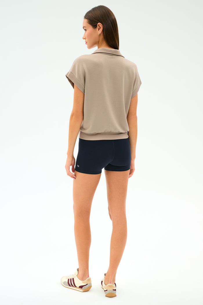 A woman stands facing away, wearing the SPLITS59 Evie Fleece Sleeveless Polo in Latte, black shorts, and beige sneakers with burgundy stripes against a plain white background.