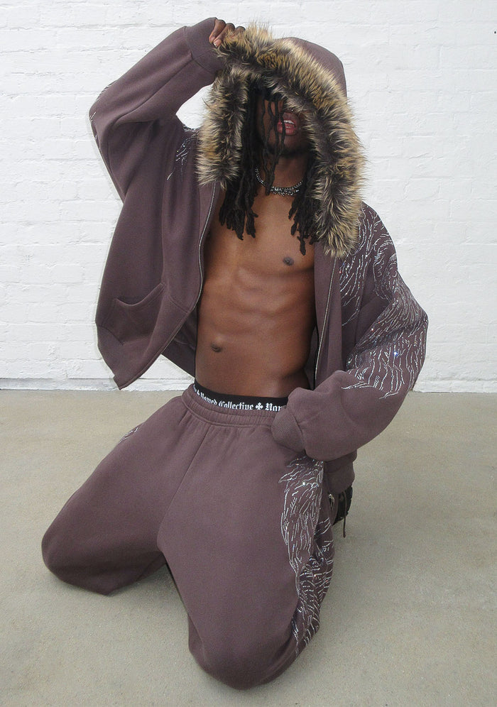 Model kneeling in brown sweatpants with rhinestone design and fur-lined hood.
