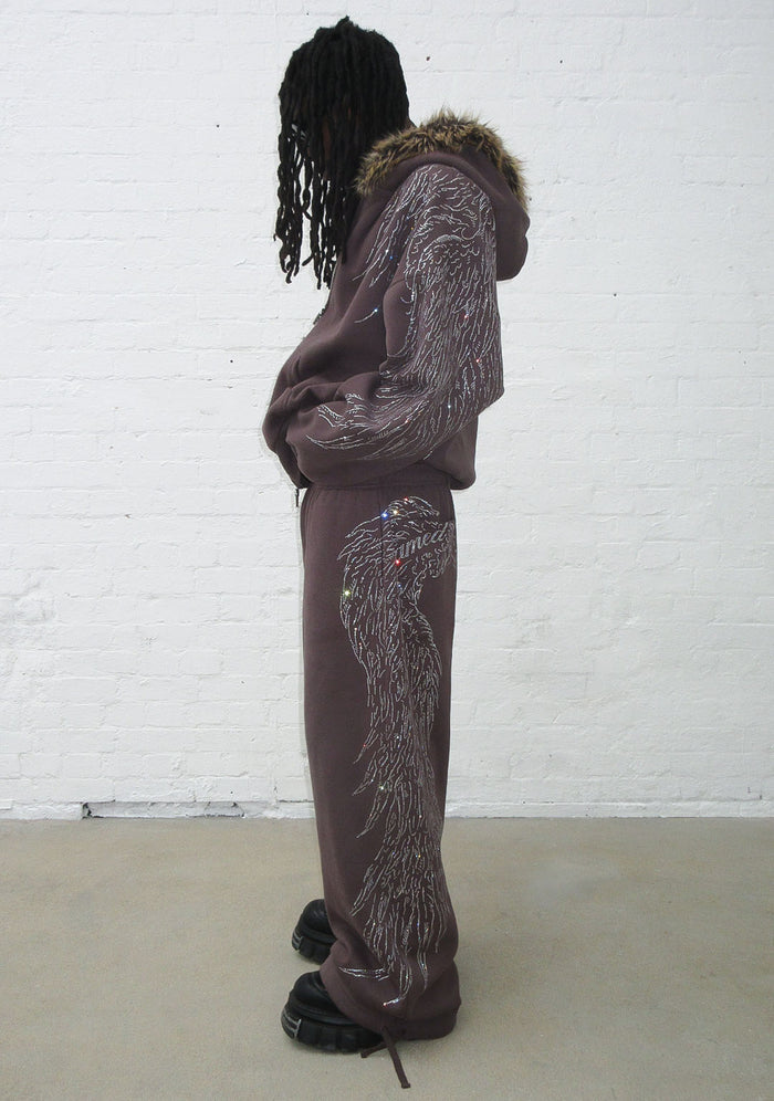 Model wearing brown sweatpants with rhinestone wing design, showing side view.