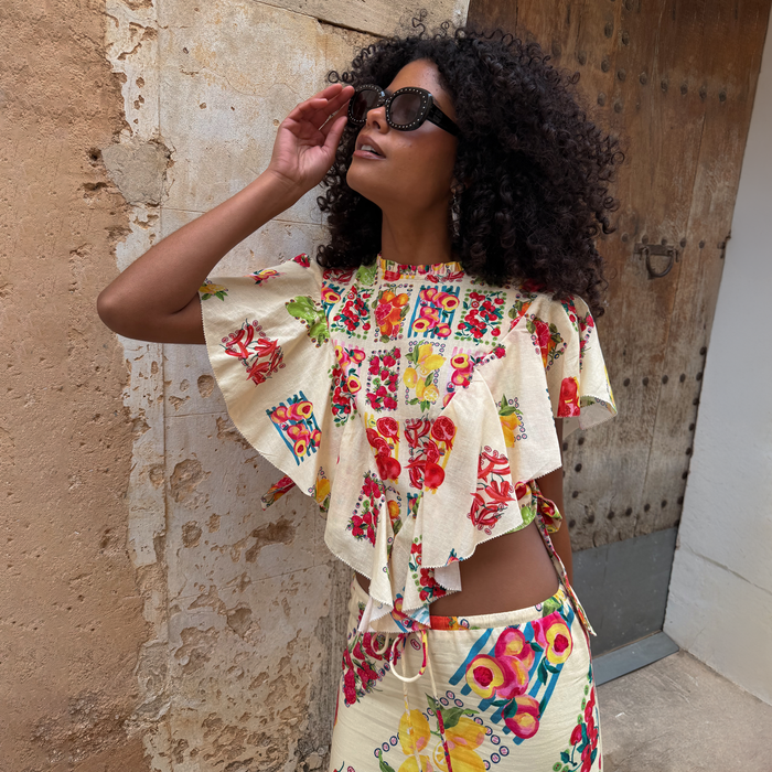Fruit Stamps Florence Top in cream cotton-linen with a Mediterranean fruit print and ruffles, on a model in a sunny setting.