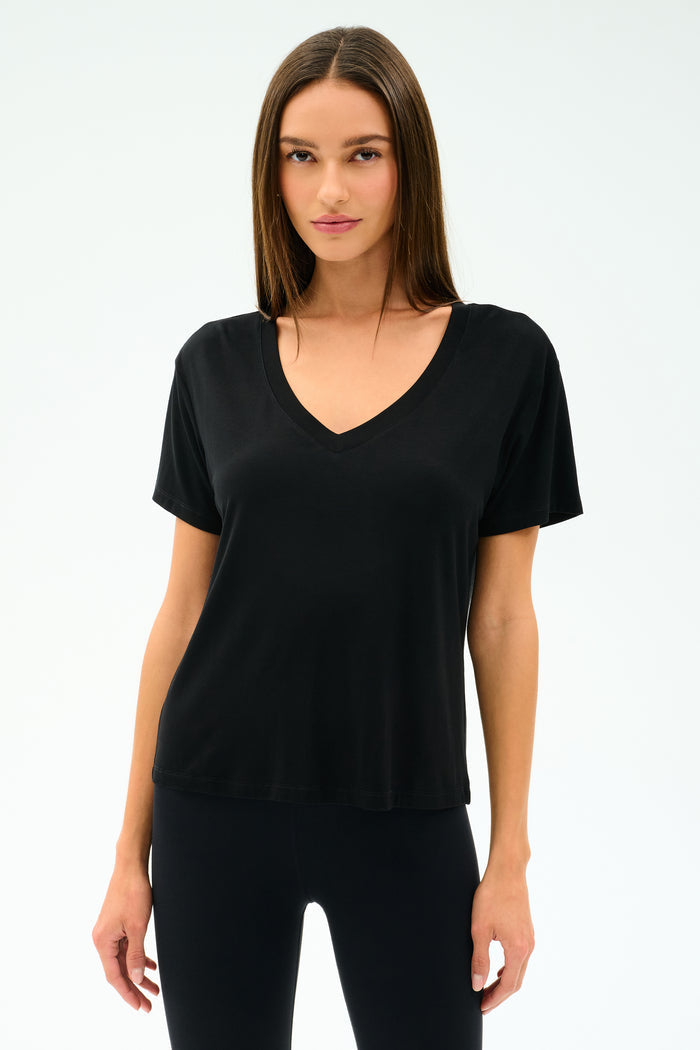 A woman faces forward, wearing the SPLITS59 Georgia Jersey V-Neck in black—crafted from a soft viscose-spandex blend—paired with black pants, set against a white background.