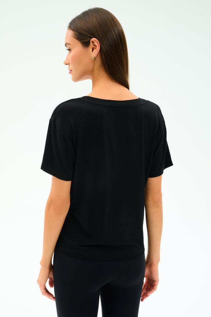 A woman with straight brown hair is standing and facing away from the camera against a plain light background, wearing the SPLITS59 Georgia Jersey V-Neck - Black and black pants.
