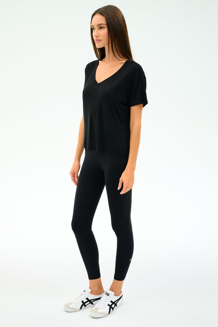 A woman stands against a plain background wearing the SPLITS59 Georgia Jersey V-Neck in black, paired with black leggings and white sneakers with black accents.