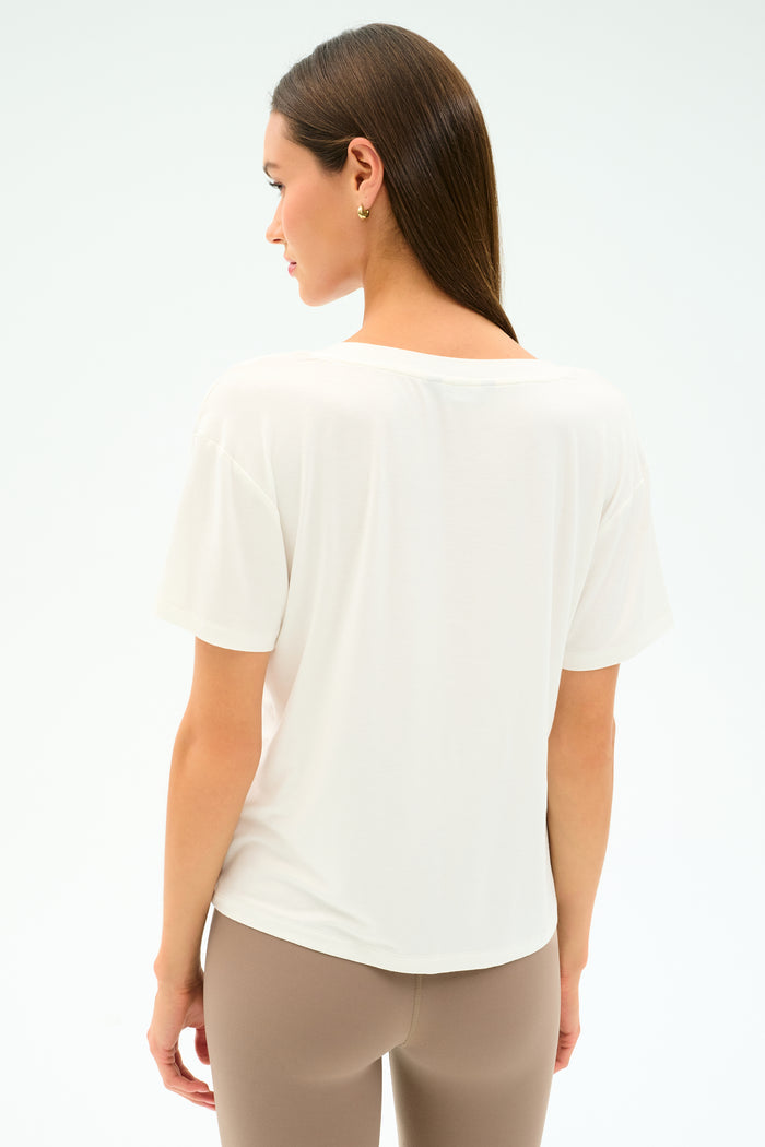 A woman stands with her back to the camera, wearing the SPLITS59 Georgia Jersey V-Neck in white and beige leggings, made from a soft viscose-spandex blend, set against a plain light background.