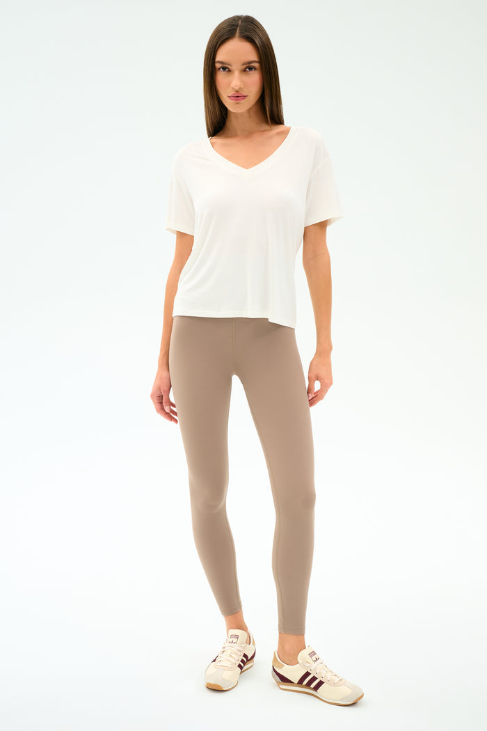 A woman faces forward against a plain white background, wearing the SPLITS59 Georgia Jersey V-Neck in white, styled with beige leggings and beige sneakers with maroon stripes.